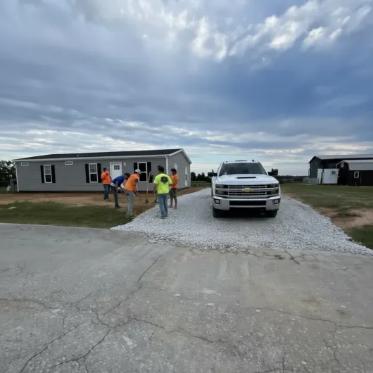 Crew laying gravel driveway in front of a gray house. White truck parked on new gravel, under cloudy sky.