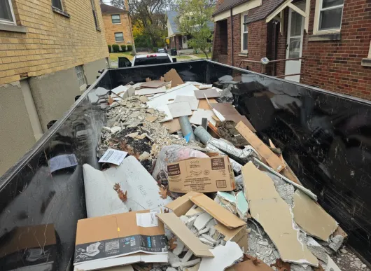 Dumpster filled with construction debris, including cardboard and drywall, in a residential alley.