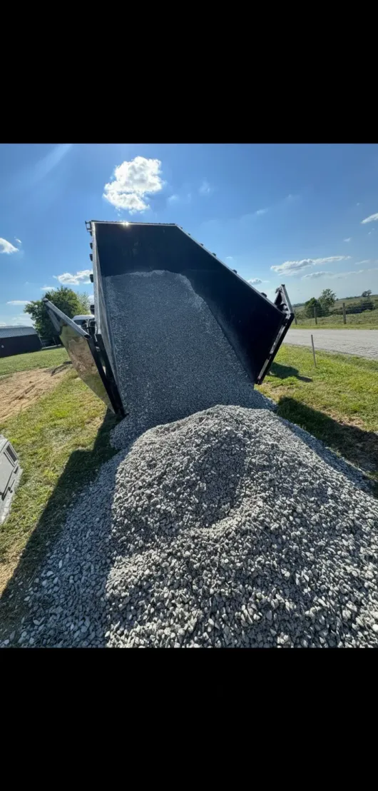 A dump truck unloading gravel on grass, bright sunlight and a blue sky in the background.