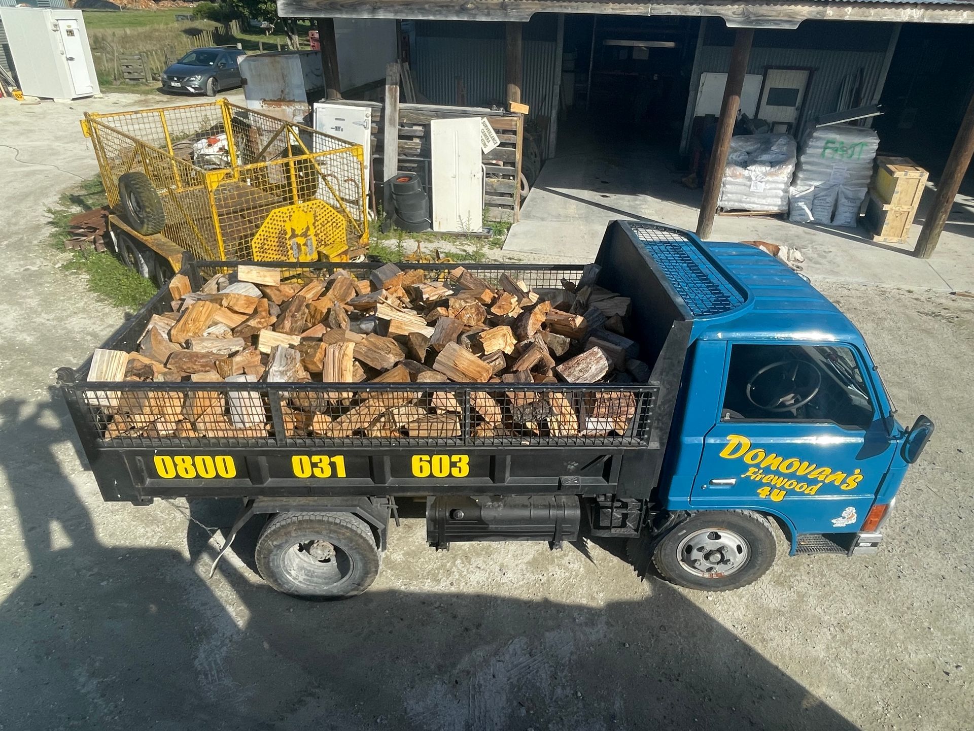 A blue dump truck filled with logs is parked in front of a building.
