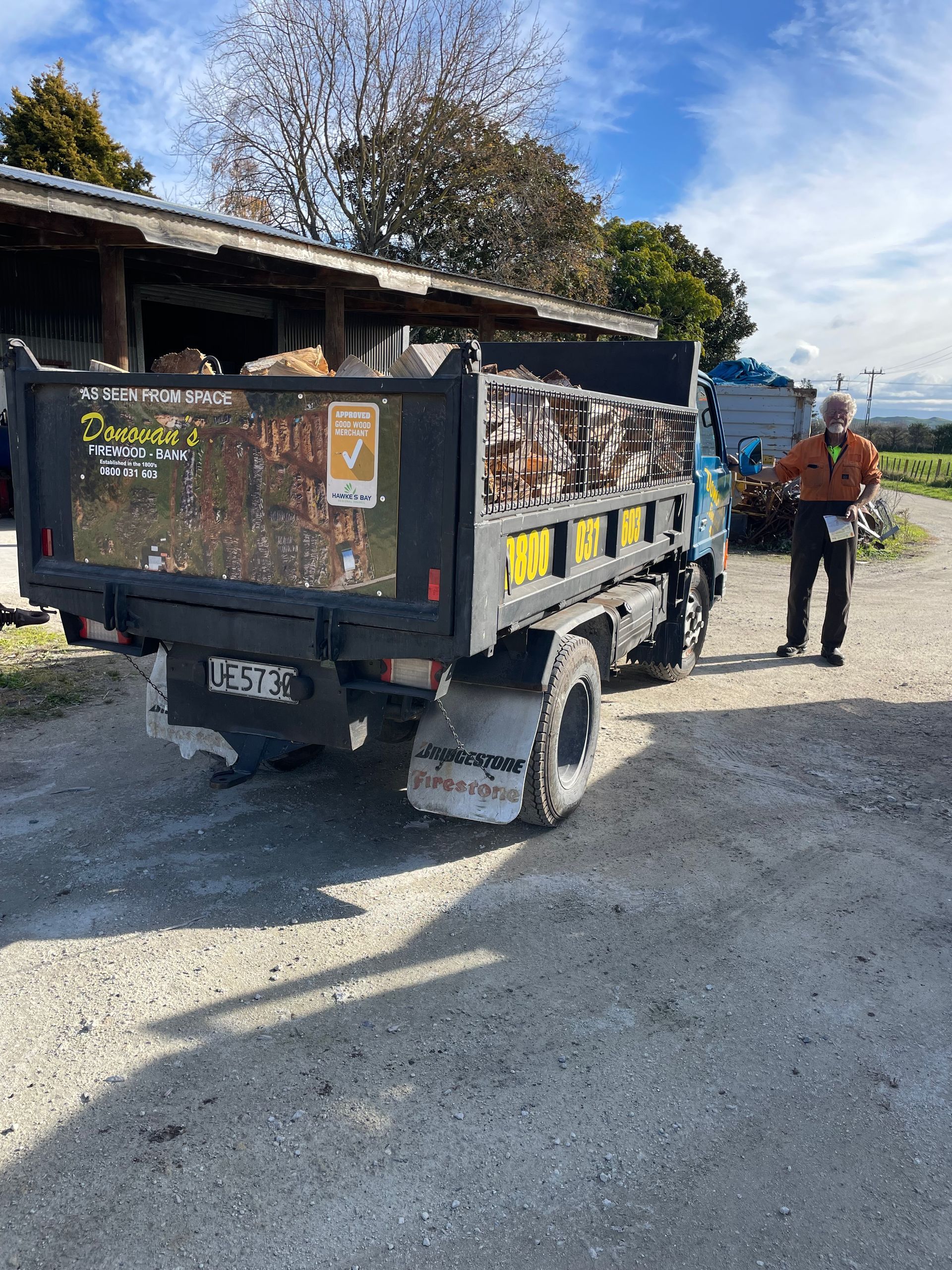 A man is standing next to a dump truck filled with wood.