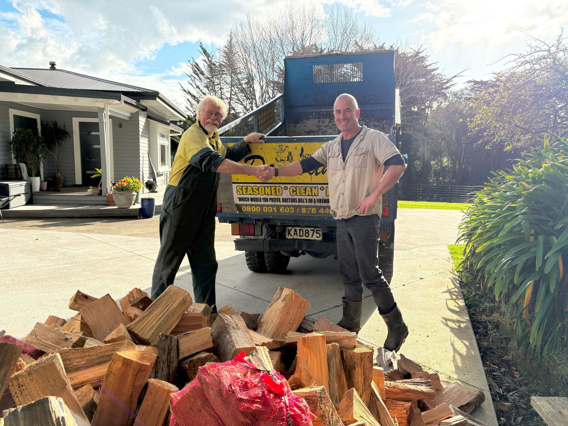 Two men shake hands in front of a truck filled with firewood in a driveway. A pile of split logs is in the foreground.