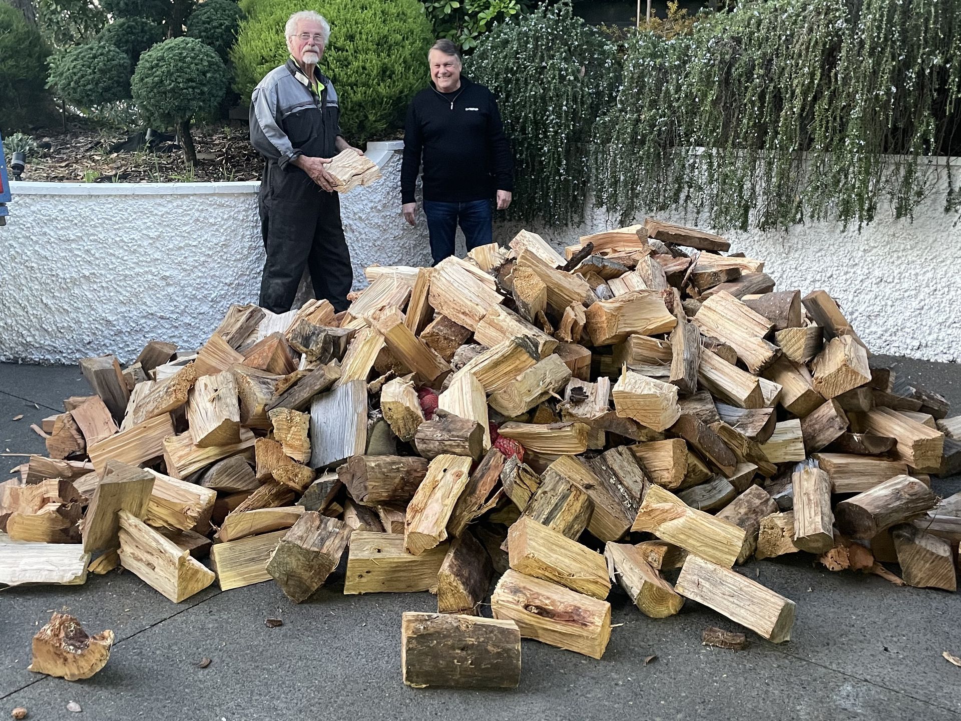 Two men standing near a large pile of chopped firewood in a driveway. One holds wood; the other smiles.