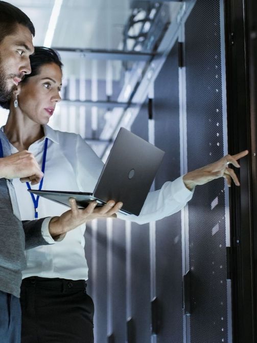 A man and a woman are looking at a laptop in a server room.