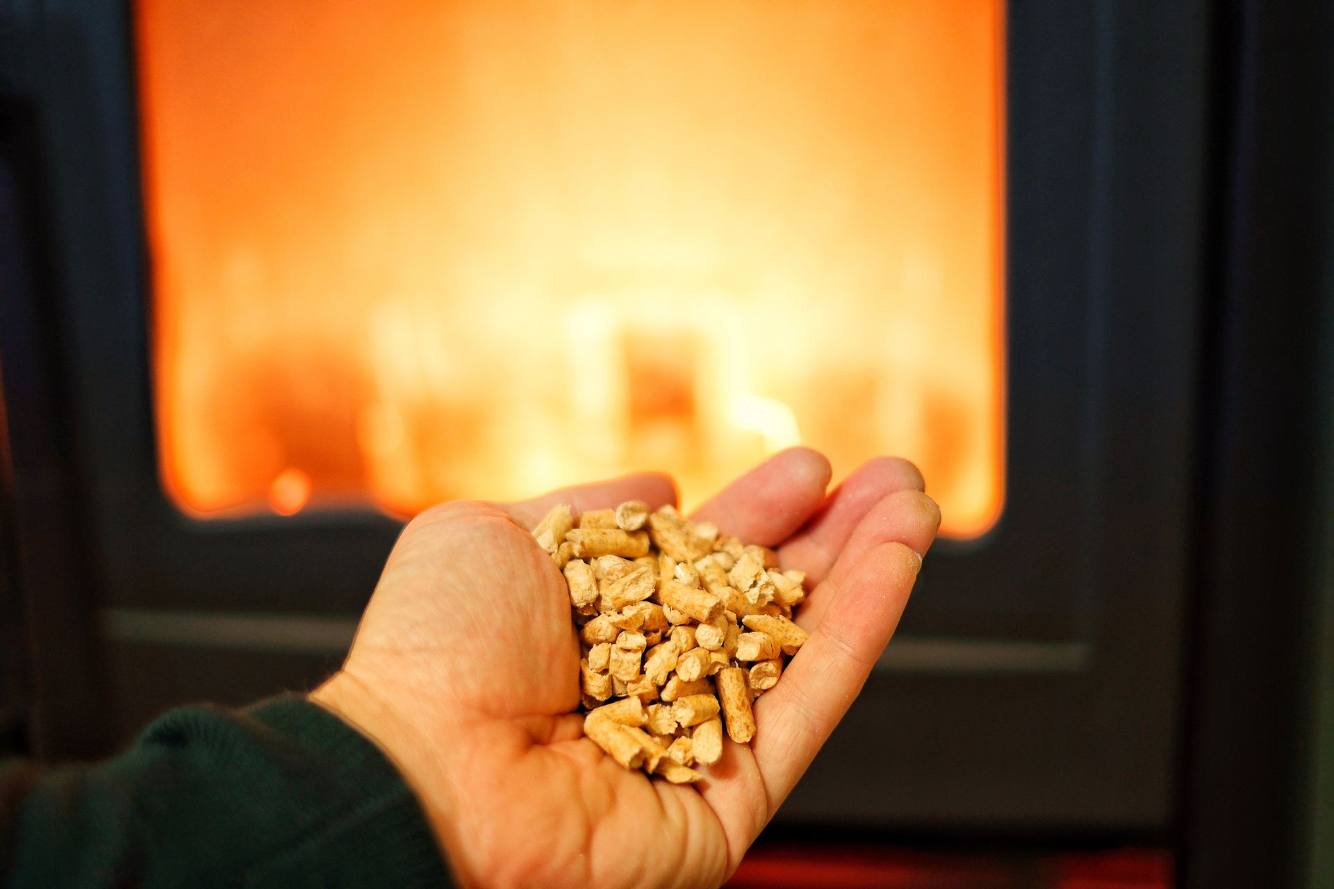 Mano che tiene pellet di legno davanti a una stufa accesa. Caldo bagliore arancione sullo sfondo.