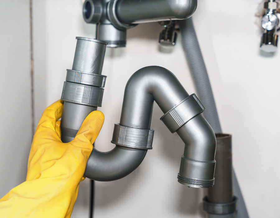 Mano in guanto giallo che tiene un tubo idraulico grigio sotto il lavello della cucina.