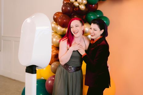 Two women laughing and posing at an iPad-powered EverGlow Booths photo booth in Boston, MA with colorful balloon backdrop