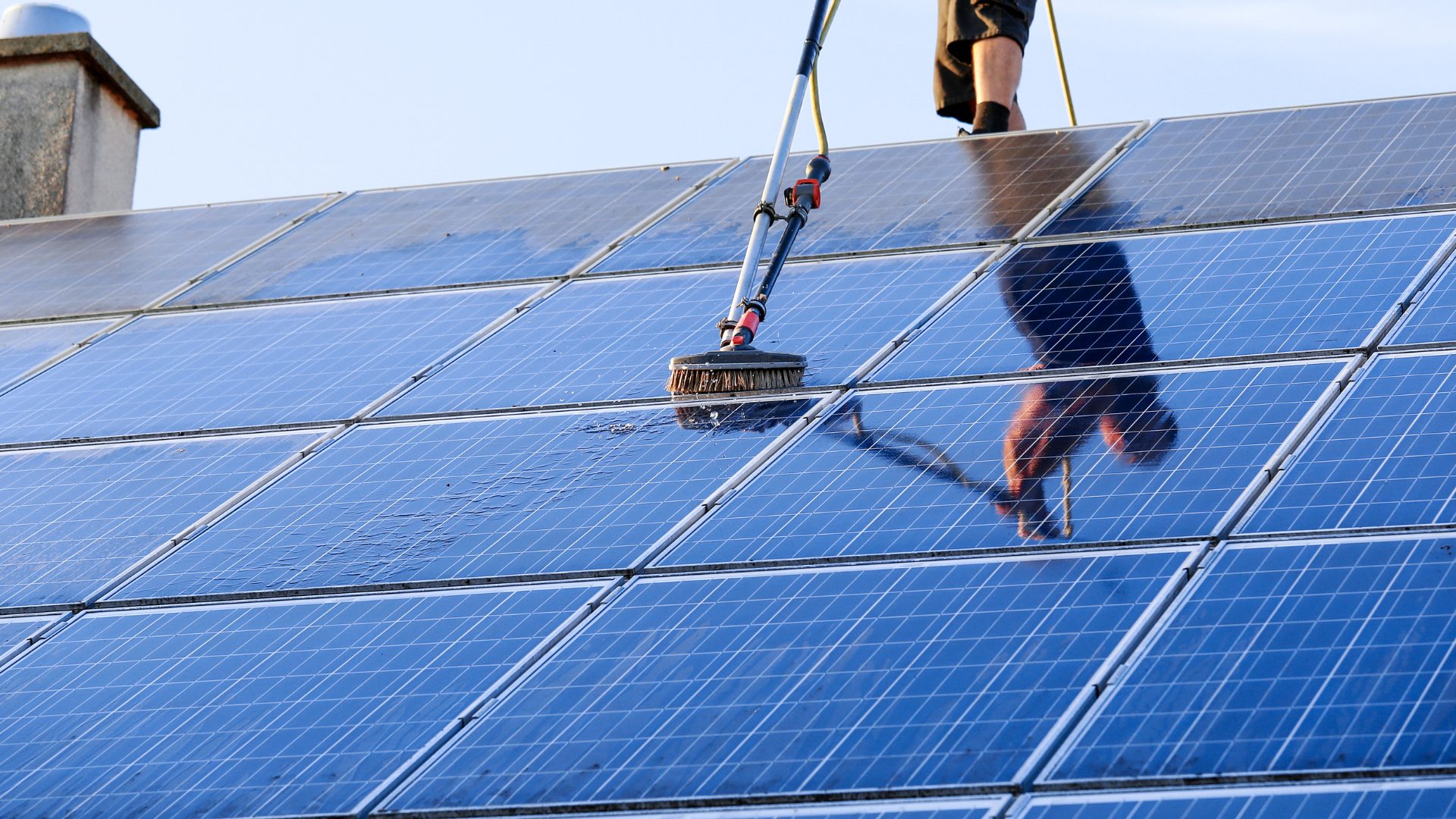 Person cleaning solar panels on a roof with a long-handled brush.