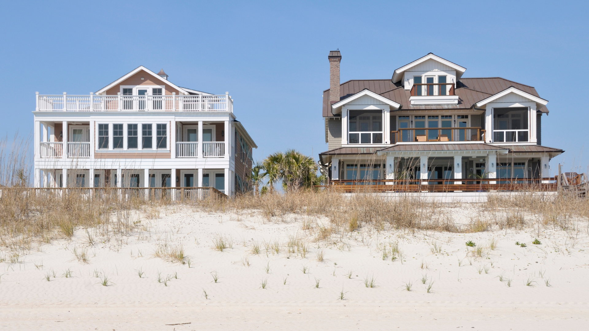 Two multi-story beach houses on sandy dunes under a clear blue sky.