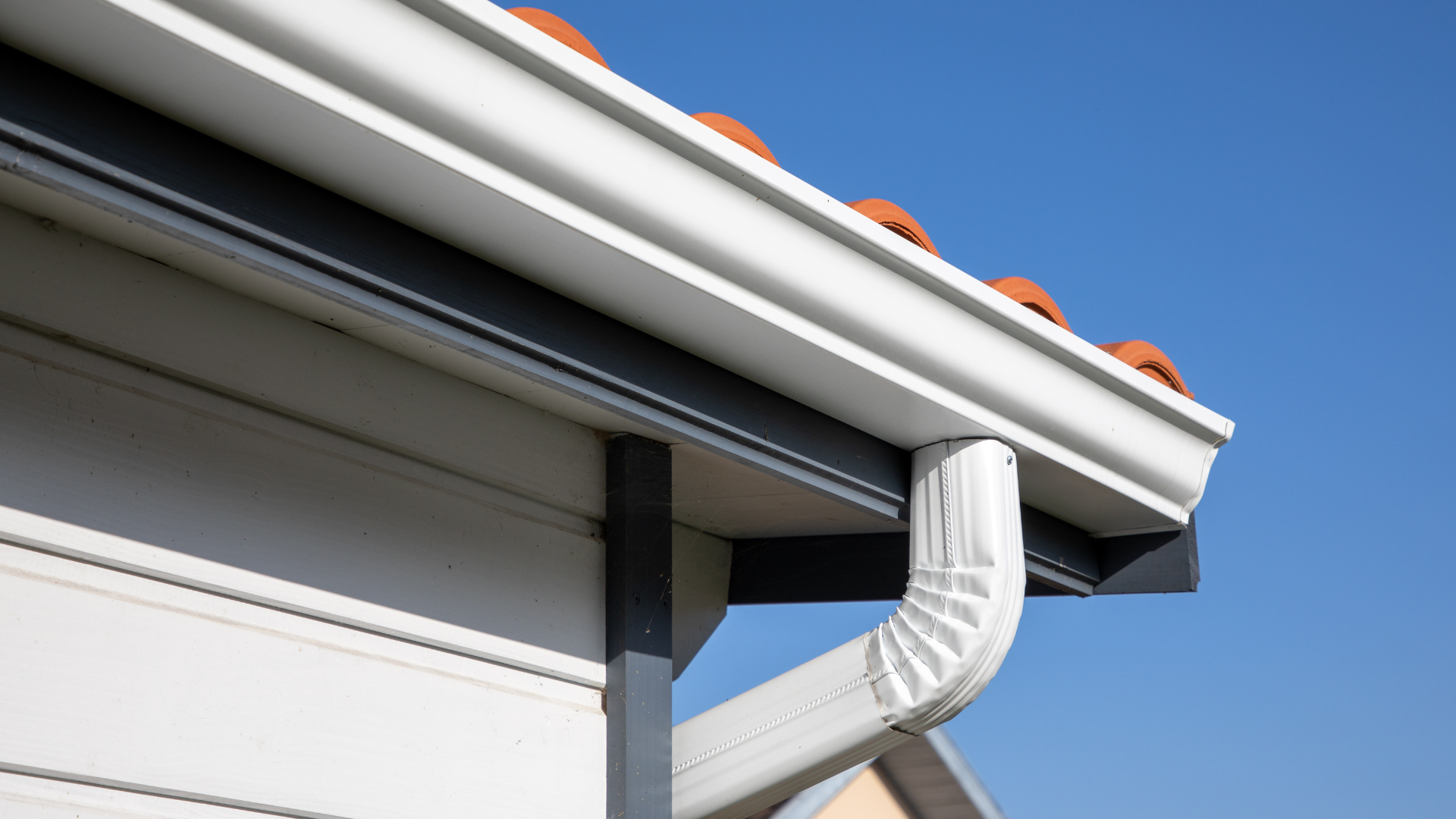 White rain gutter with downspout on a house with an orange tile roof against a blue sky.