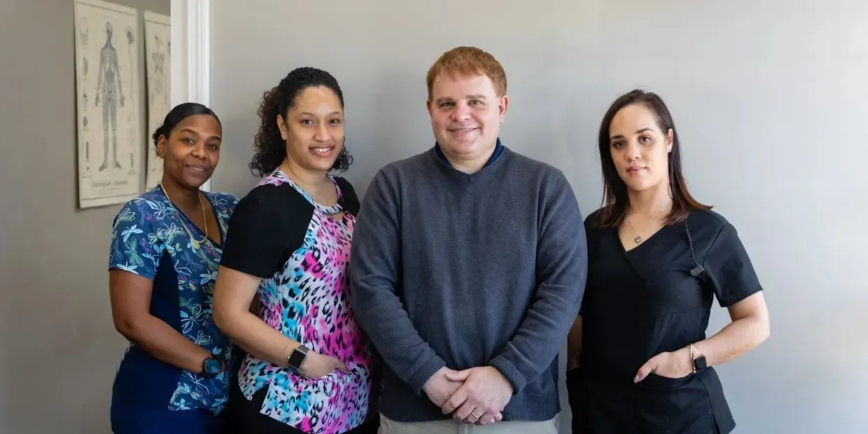 Four people standing close together in front of a light gray wall. Two are wearing scrubs, one a sweater.
