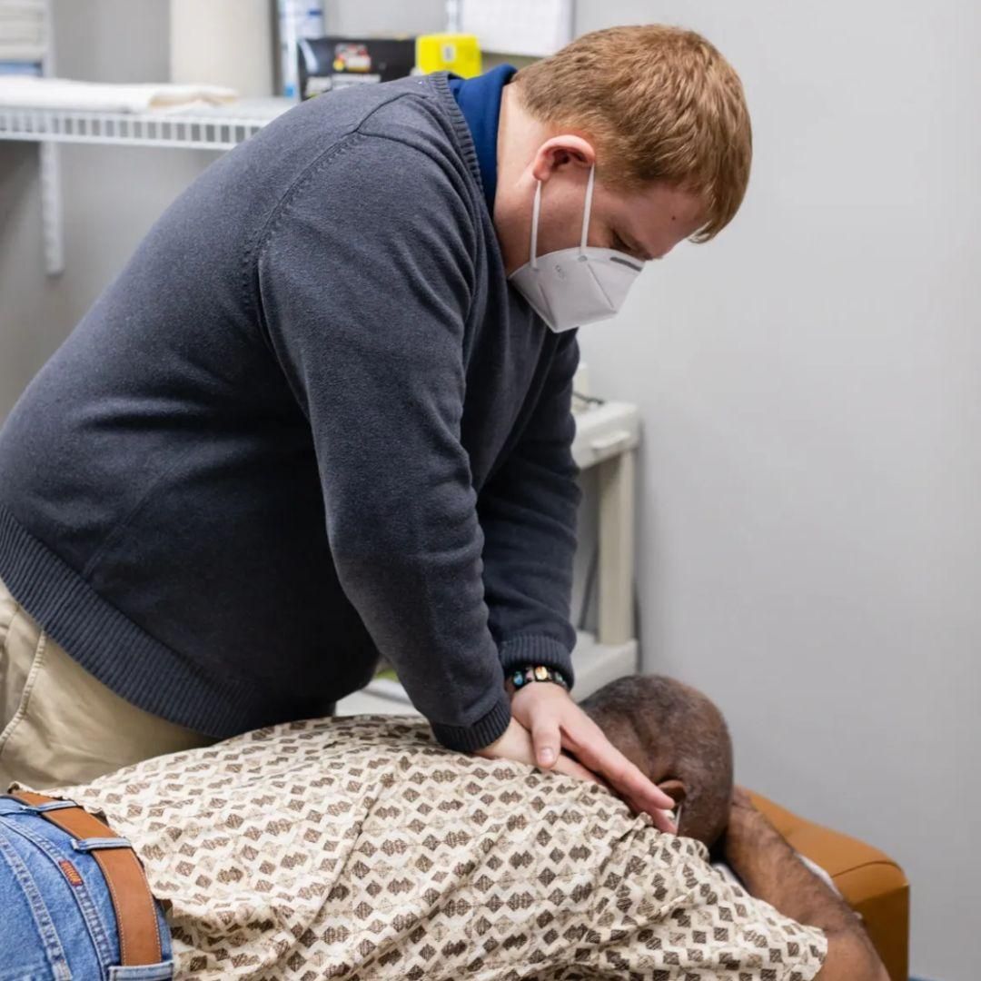 Man in mask adjusting patient's back on exam table; indoors.