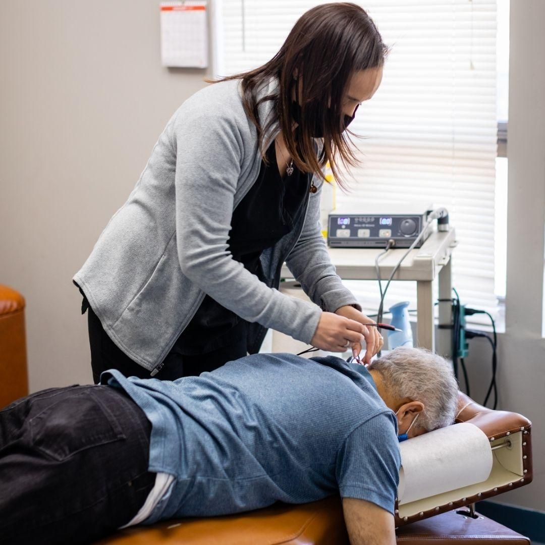 Woman in a gray cardigan adjusts wires on a person’s back.