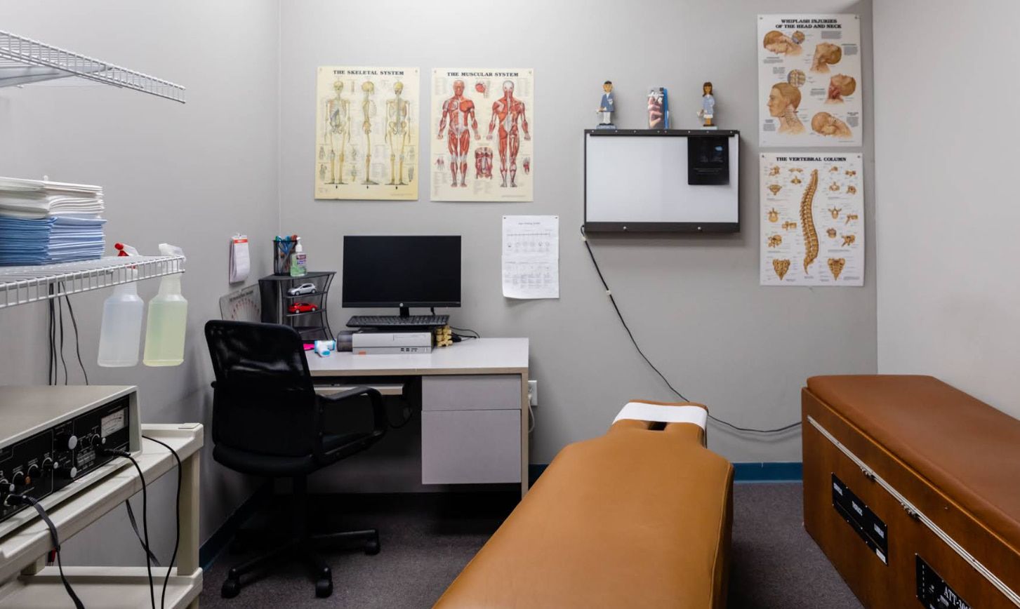 Chiropractor's office: examination table, desk with computer, anatomical charts on walls.