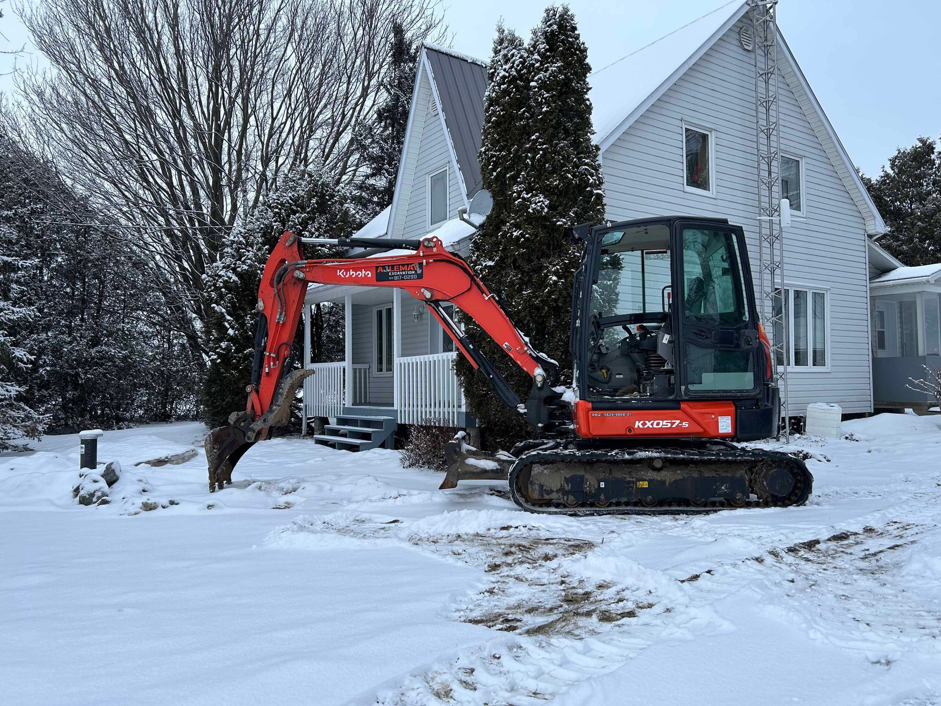Une excavatrice rouge est garée devant une maison blanche dans la neige.