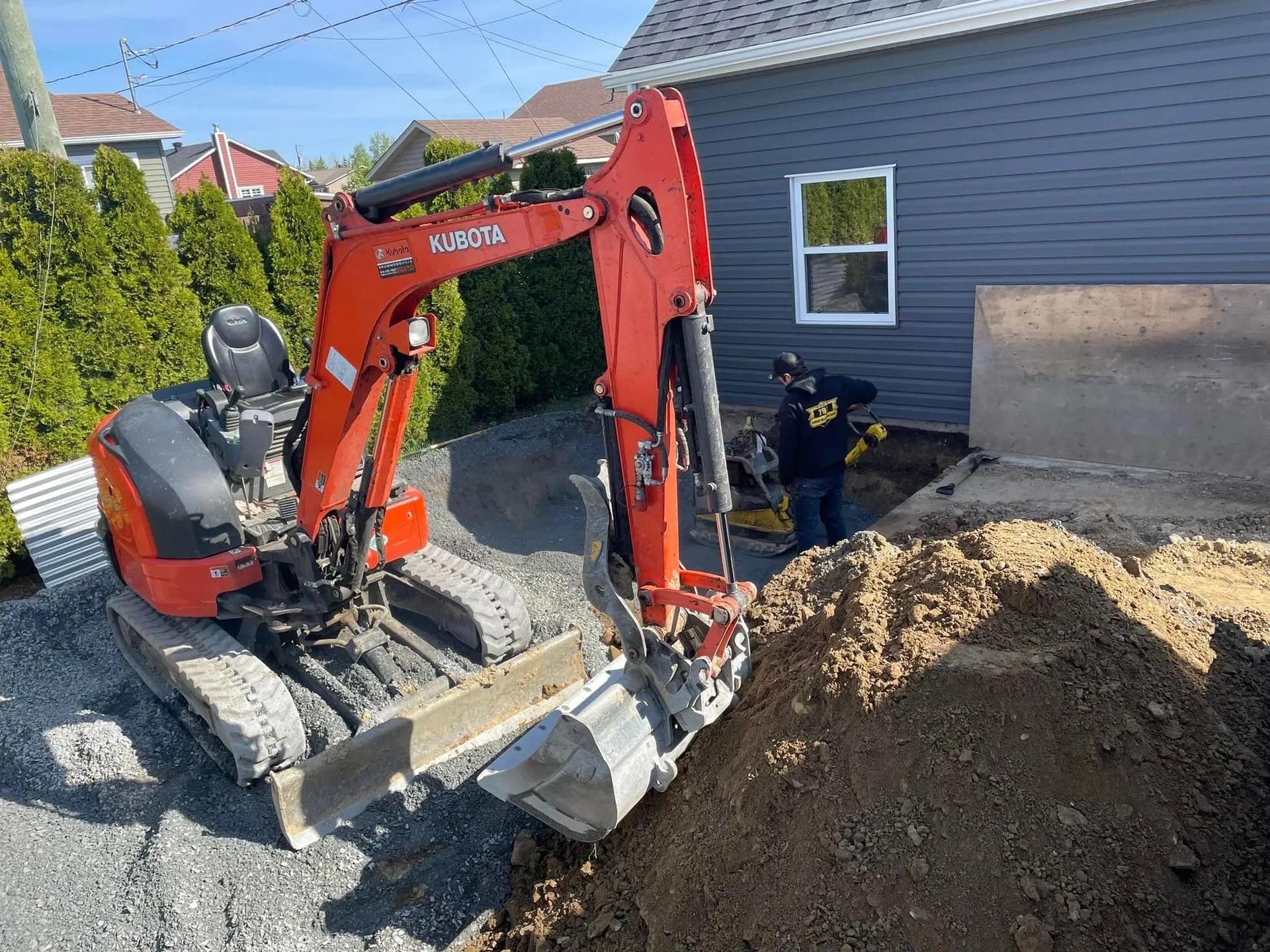 Une petite excavatrice rouge creuse un trou devant une maison.