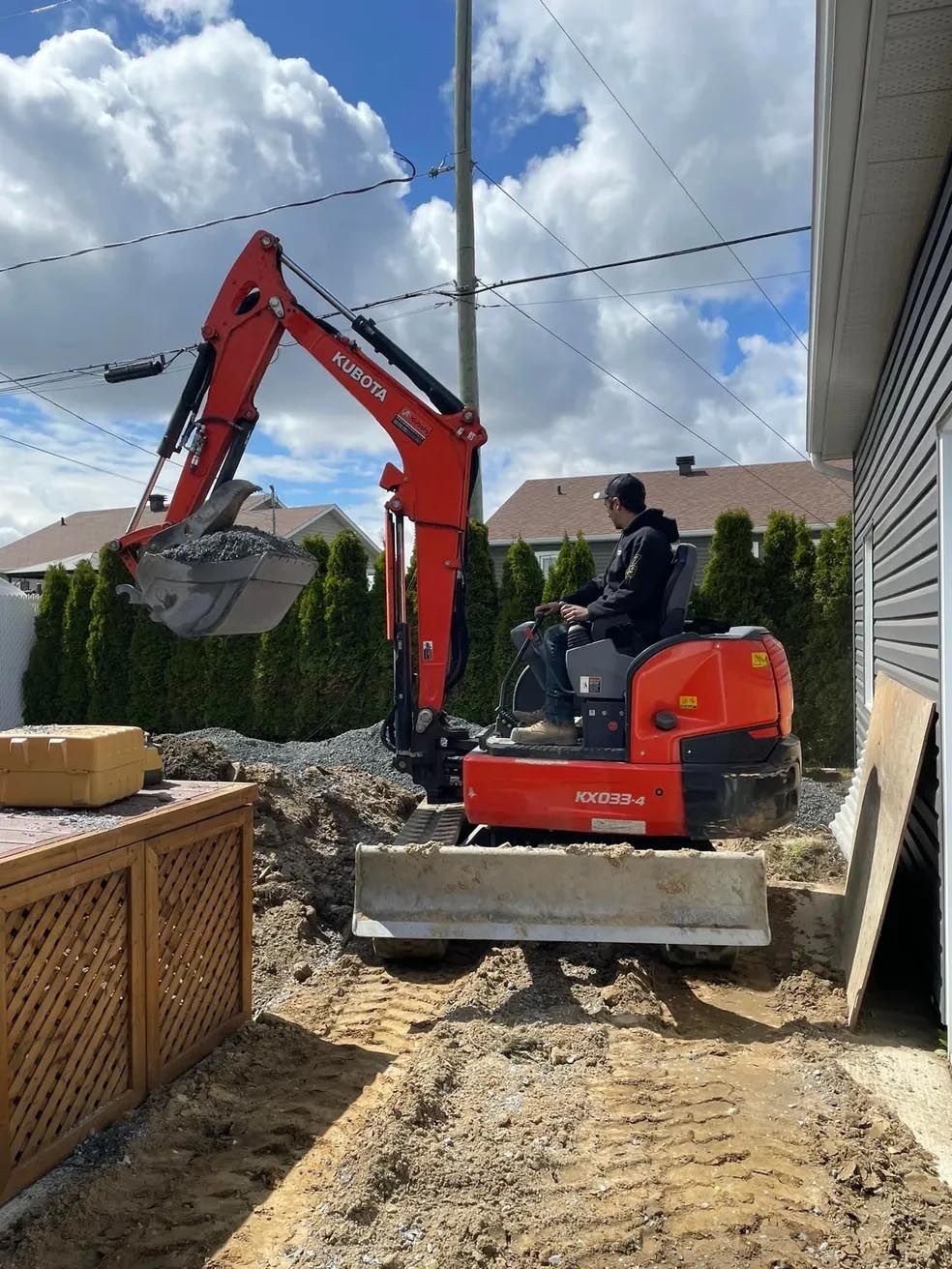 Un homme conduit une excavatrice sur un chemin de terre devant une maison.