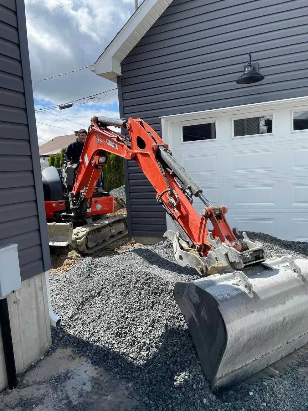 Une excavatrice est assise devant un garage à côté d'une maison.