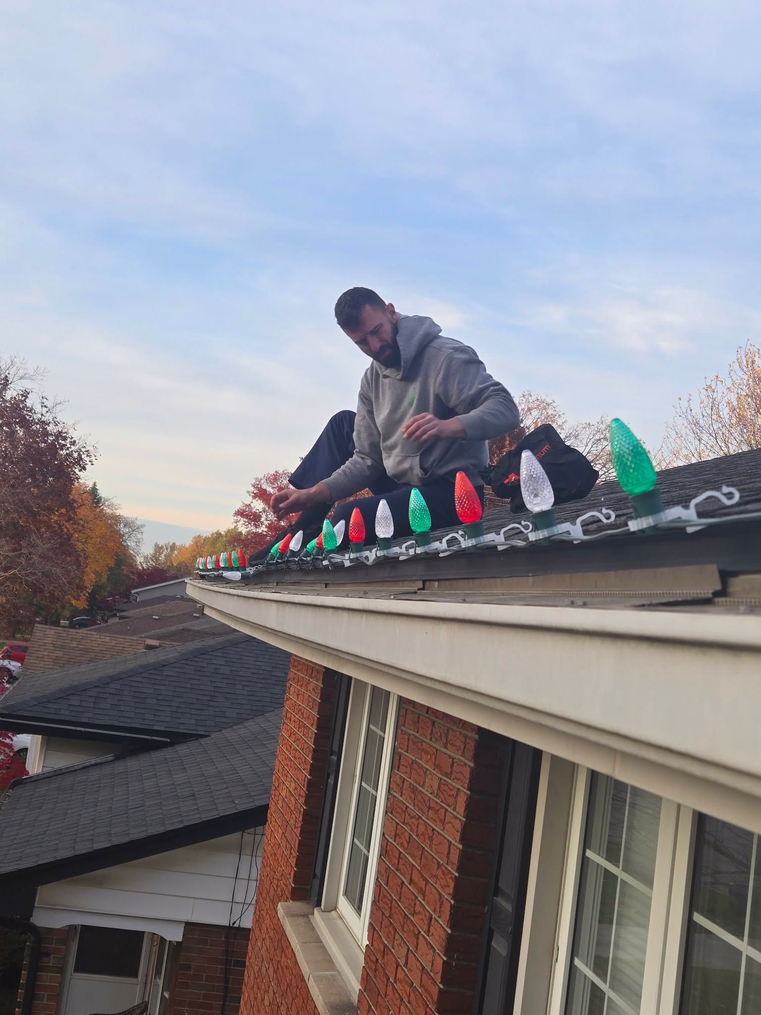 Person on a roof installing holiday lights. Red brick building with visible windows. Overcast sky.