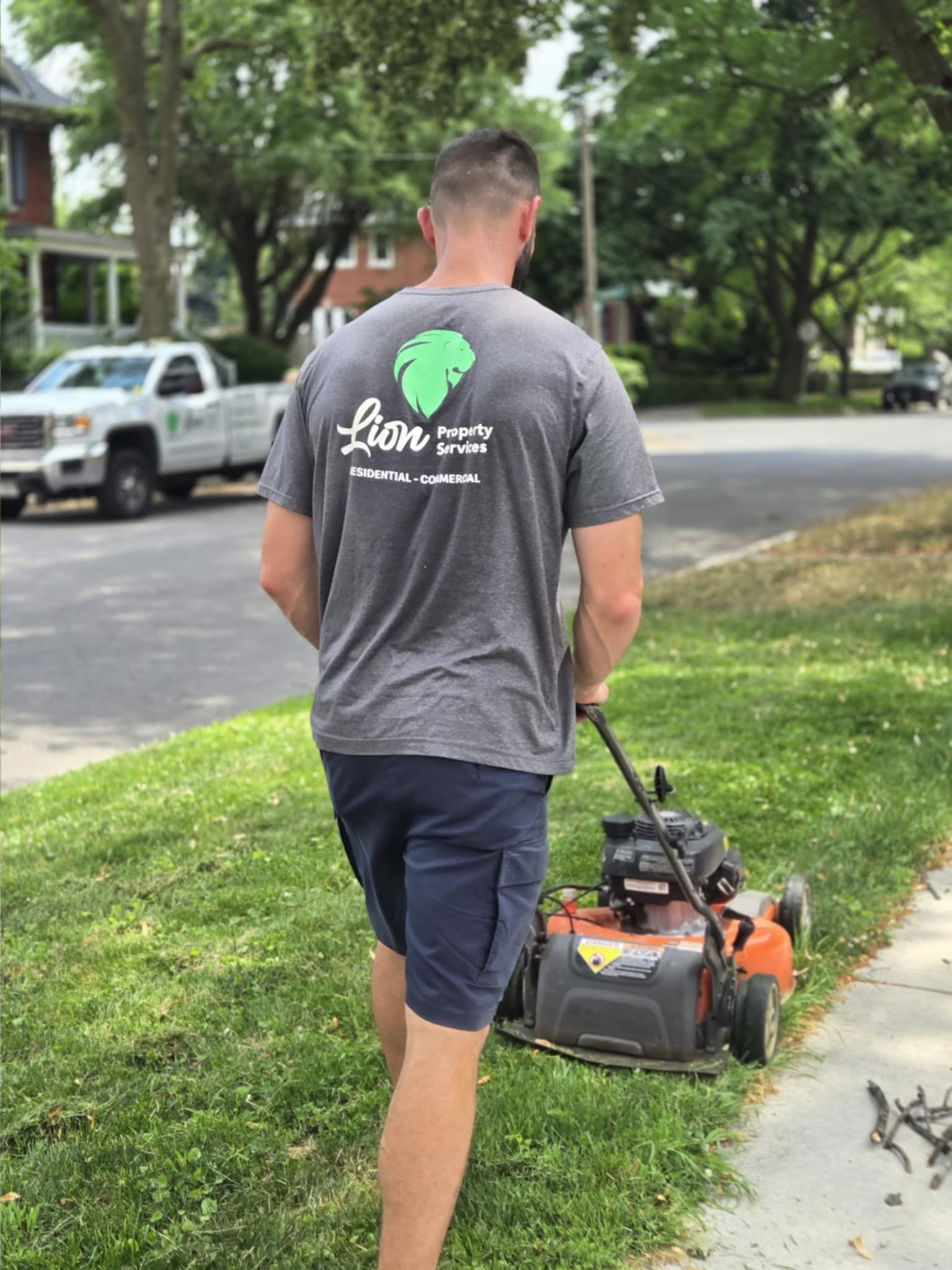 Man mowing a lawn; gray shirt with company logo, navy shorts, street and truck in background.