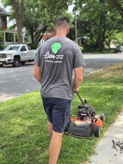Man mowing a lawn; gray shirt with company logo, navy shorts, street and truck in background.