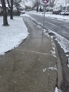 Snowy sidewalk next to a street with cars and houses; a no-entry sign is visible.