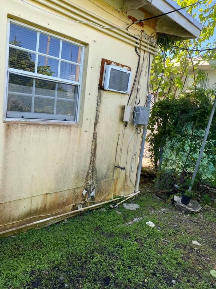 Yellow building exterior with a window, air conditioning unit, and electrical conduit, with overgrown vegetation nearby.