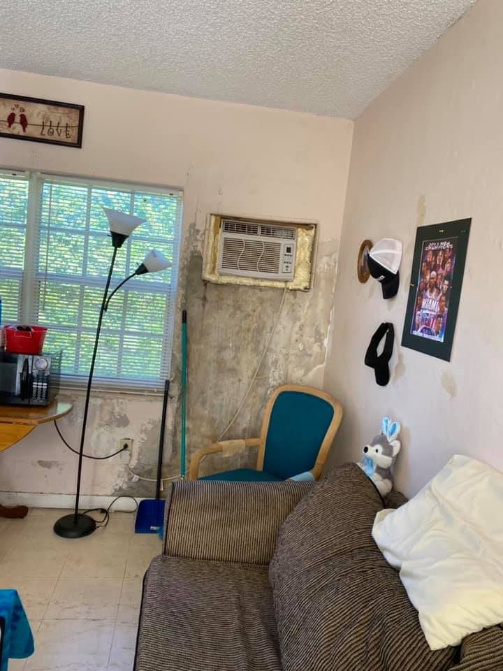 Room with window, AC unit, and worn walls. Sofa, chair, and floor lamp are present.