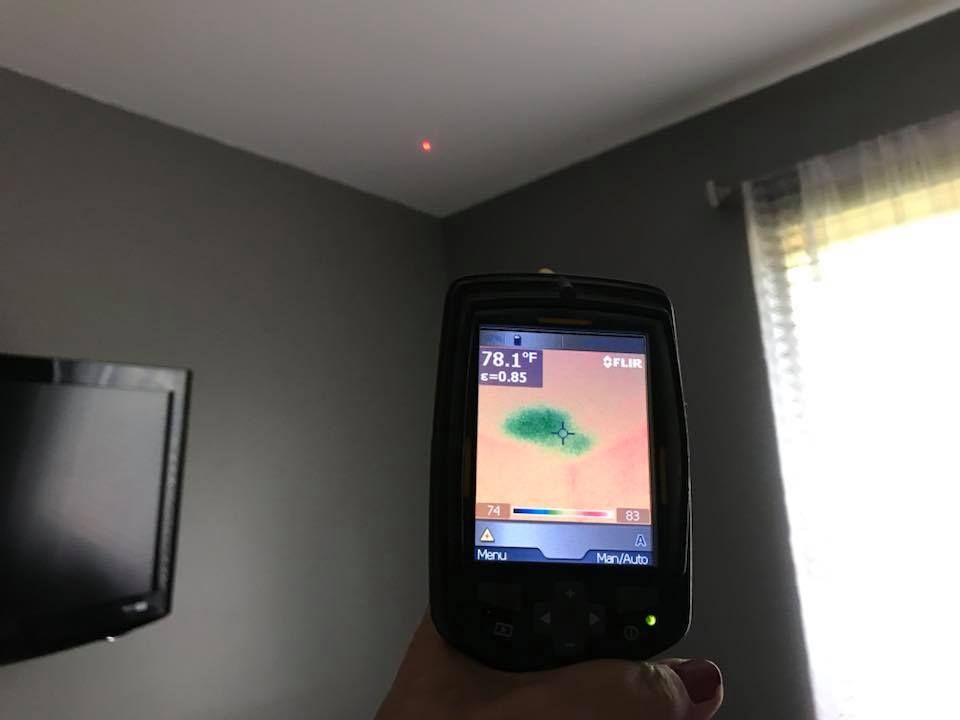 A person uses a thermal imaging camera to scan a ceiling. The camera displays a
