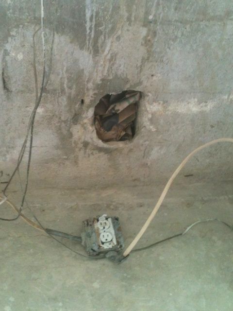 A weathered concrete surface with an electrical outlet and a damaged hole, surrounded by exposed wires.