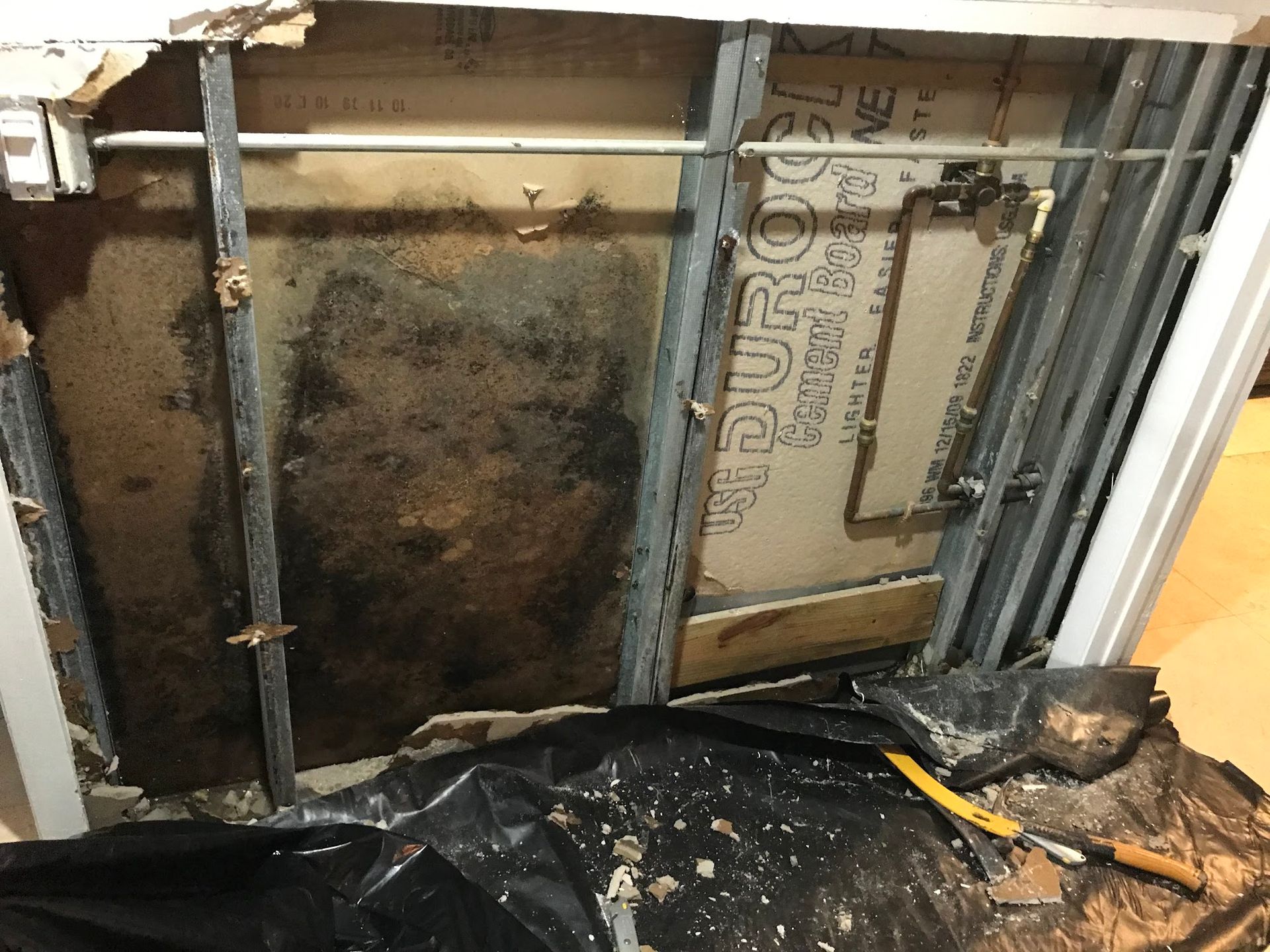 Wall damage revealing black mold, metal studs, and drywall debris. Black tarp and tools on floor.