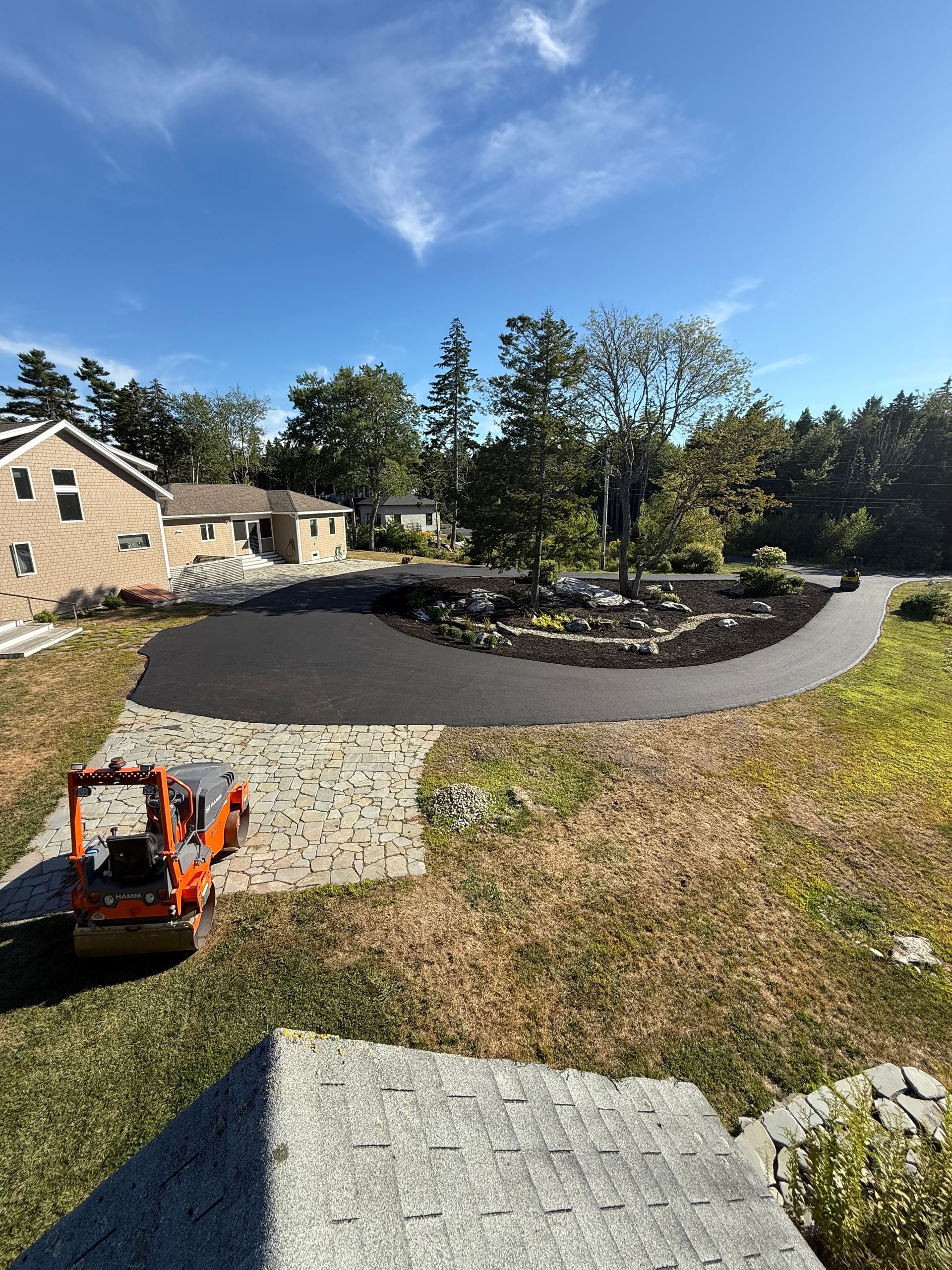 Asphalt driveway curves through a yard with a small excavator. Two houses and trees visible under a blue sky.