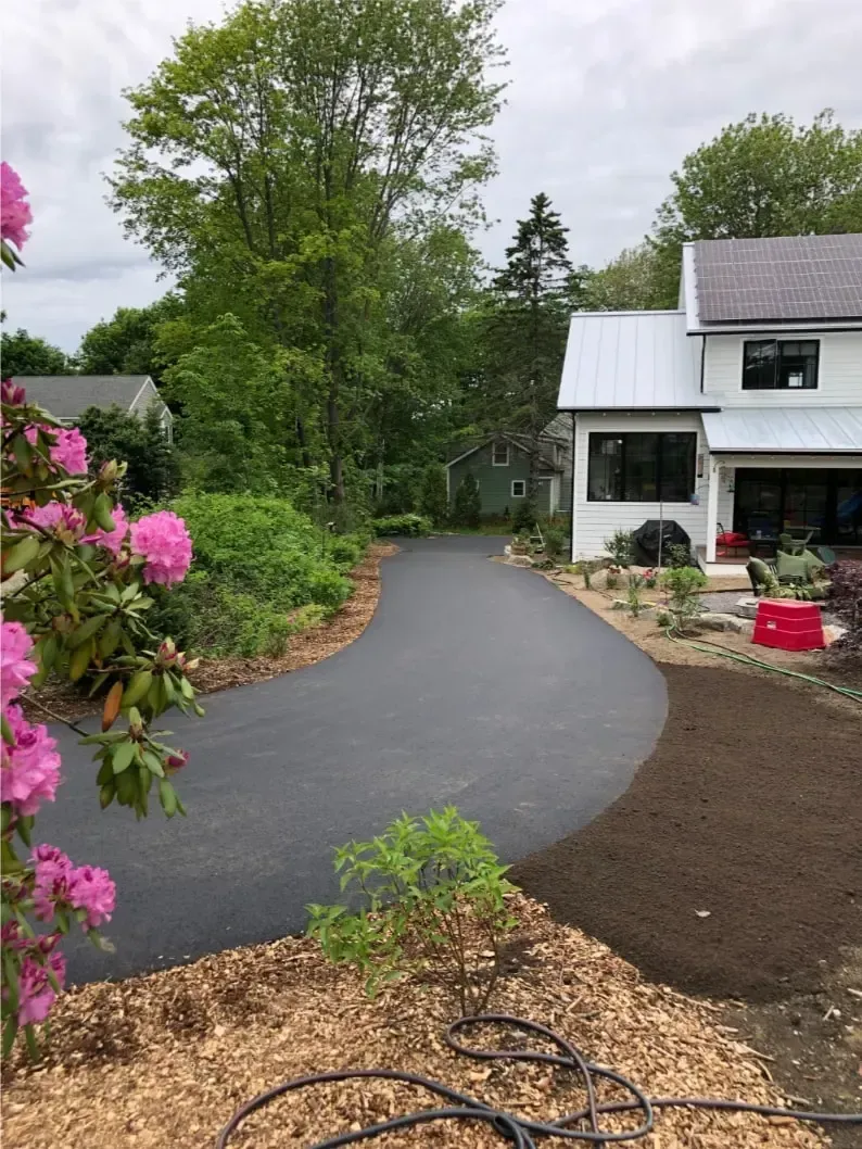 Asphalt driveway curves toward a white house with landscaping. Pink flowers and mulch border.