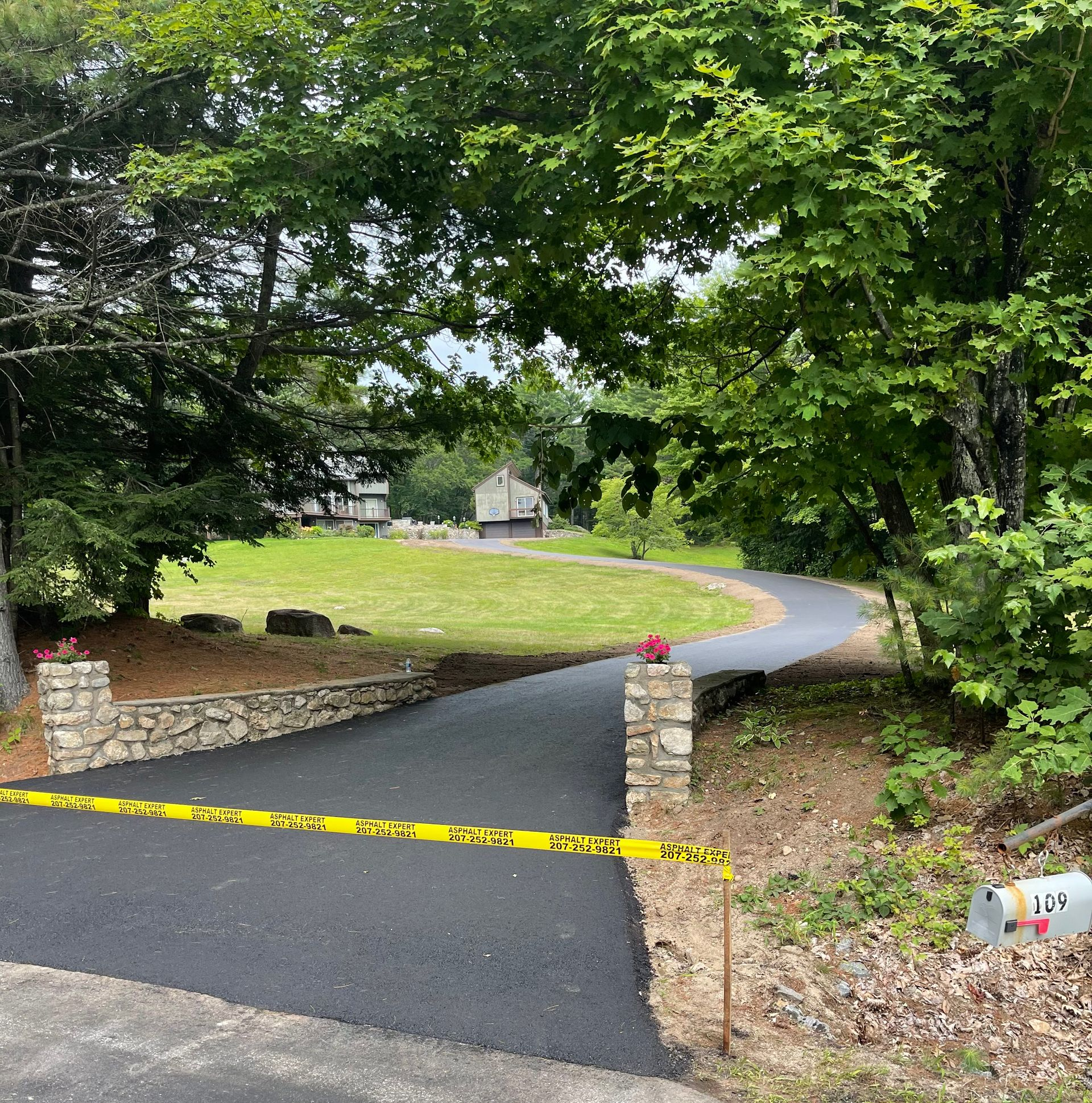 Paved Maine driveway curving into a grassy yard, flanked by trees and stone pillars with red flowers. 
