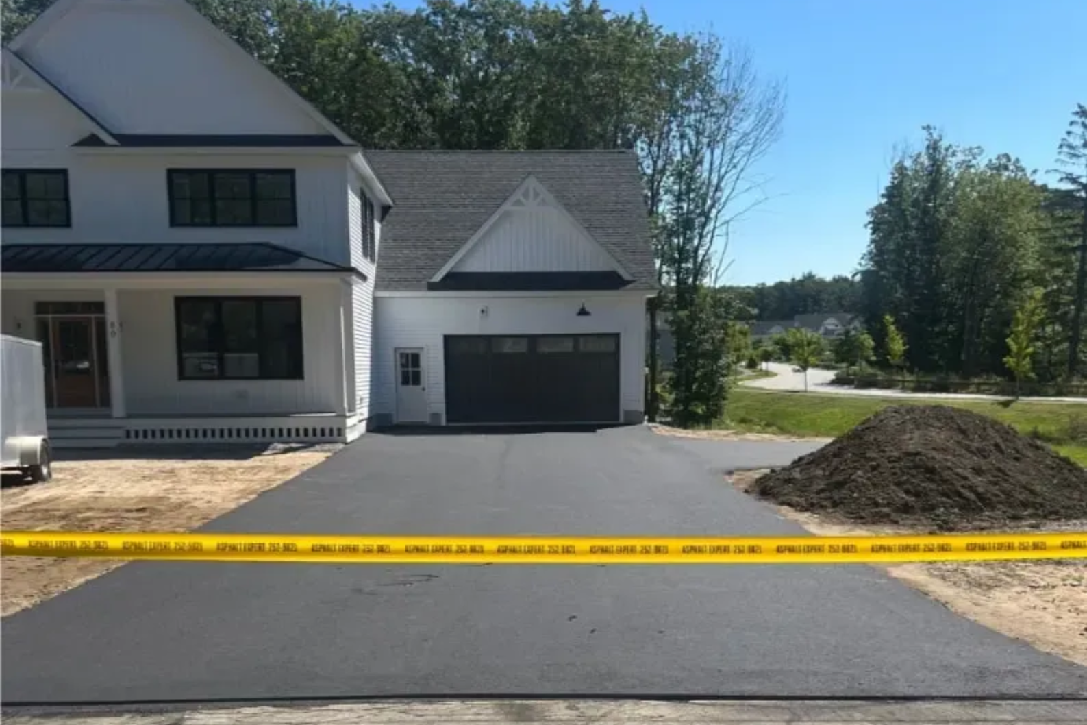 Newly paved asphalt driveway in front of a white house with a garage; yellow caution tape in foreground.