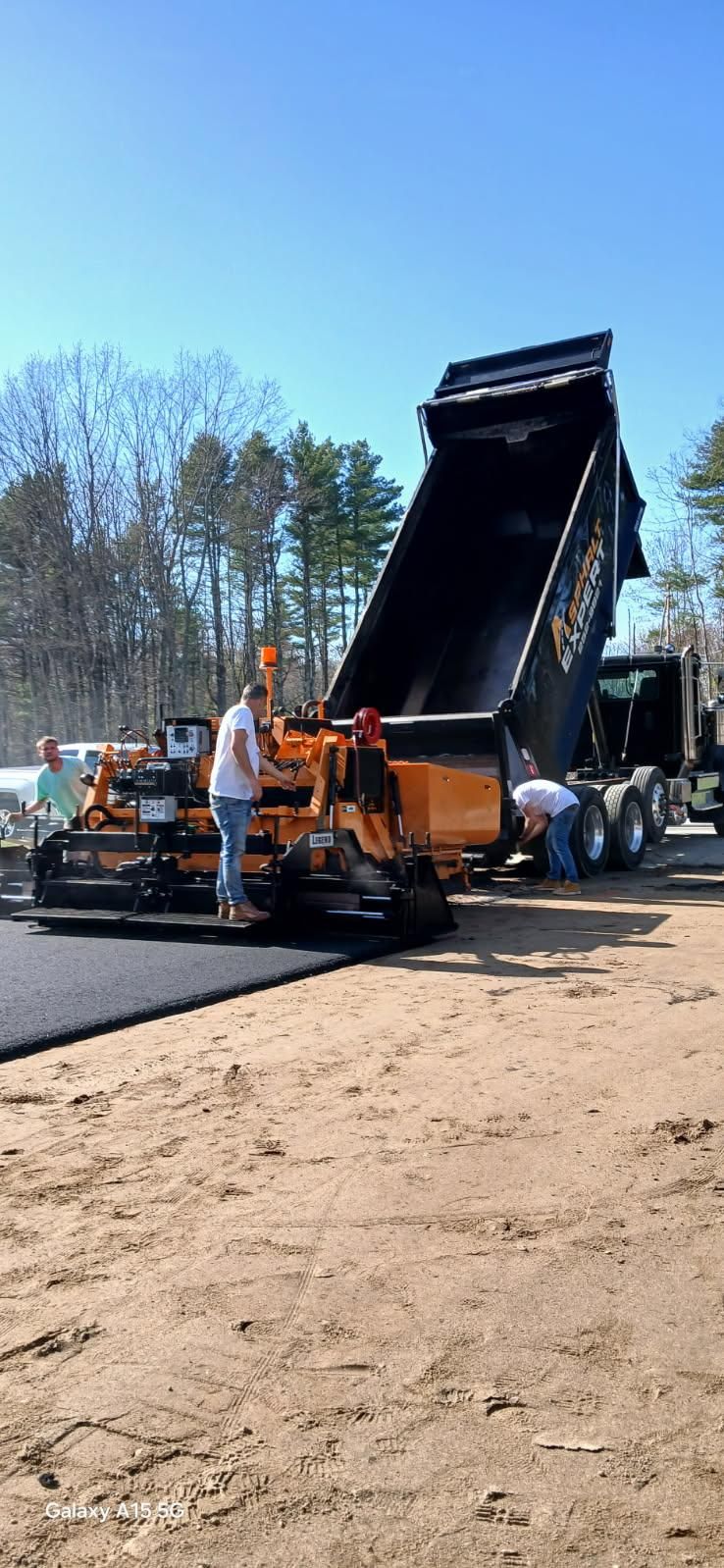 Asphalt paving crew at work in Maine, a dump truck unloading into the paver, clear blue sky.