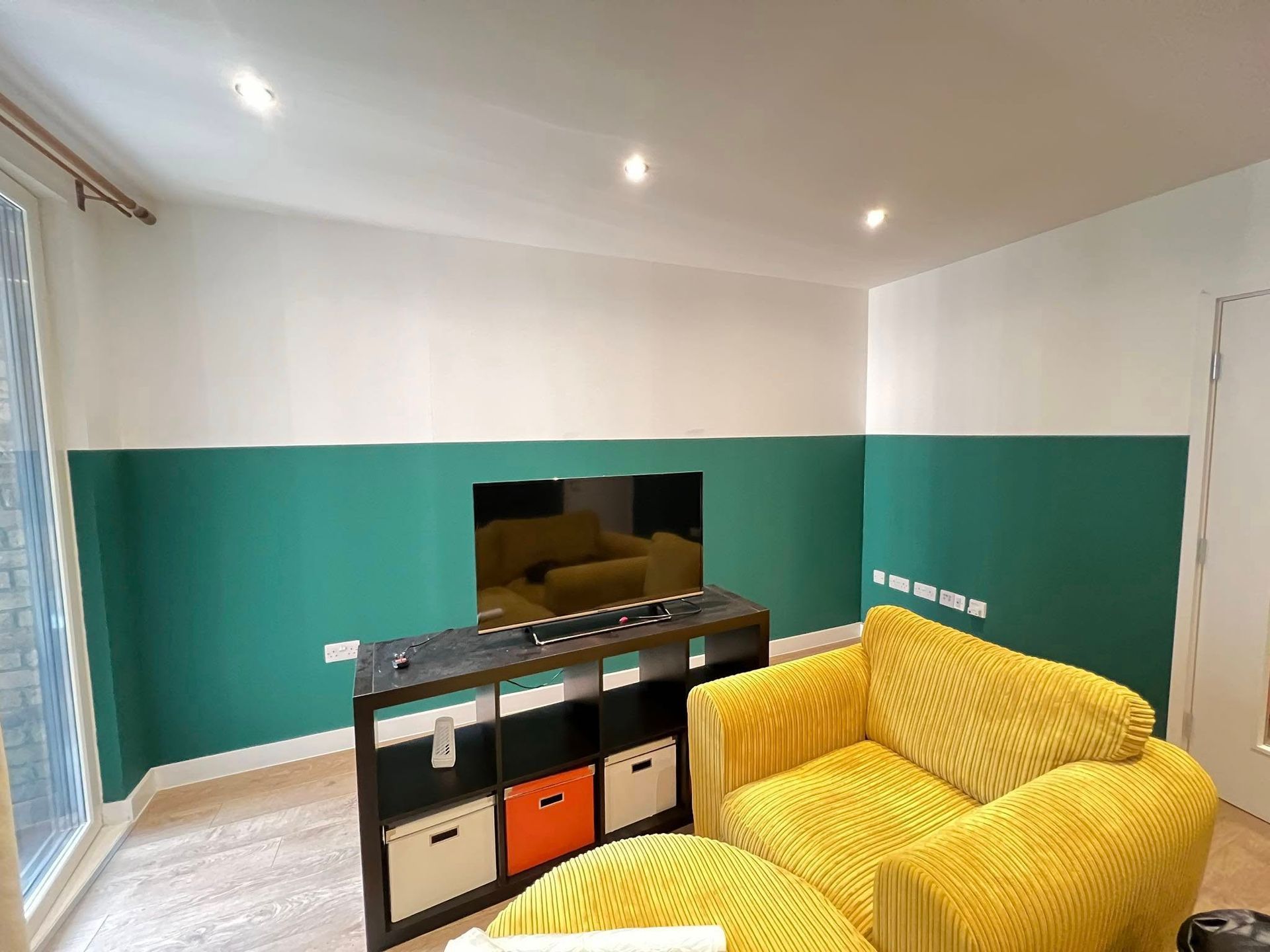 Green and white painted living room with a yellow armchair, TV, and storage unit.