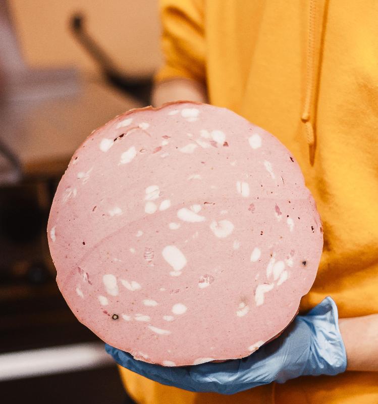 Una persona con guanti blu e camicia gialla tiene in mano una grossa fetta rotonda di mortadella.