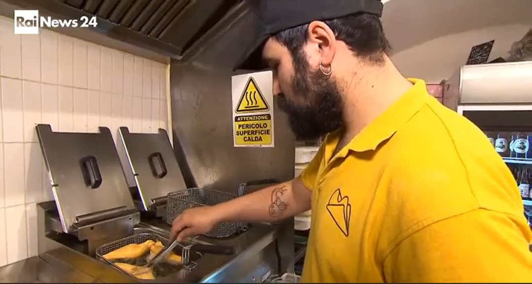 Un lavoratore con una maglietta gialla frigge del cibo in una cucina professionale, con un cartello di avvertimento giallo