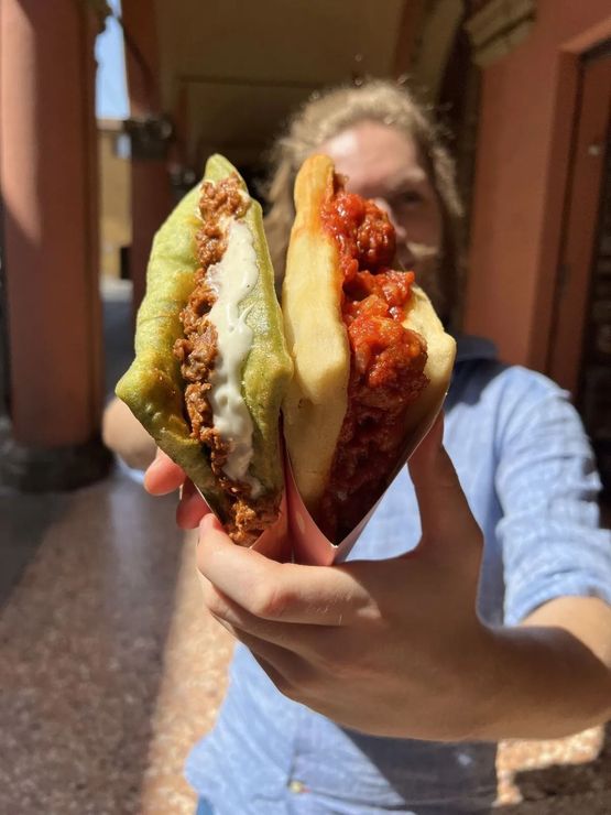 In un corridoio, una persona tiene in mano due panini fritti, uno con carne e formaggio, l'altro con salsa di pomodoro.