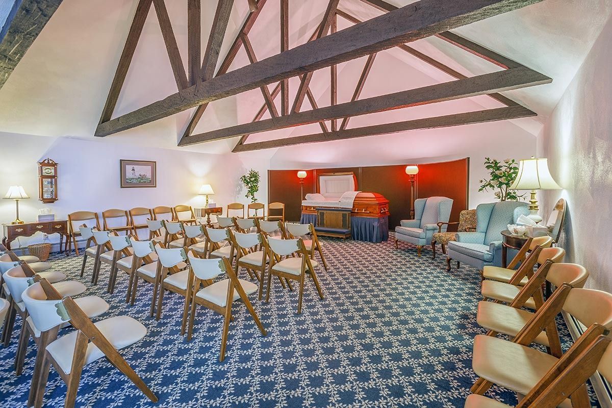 Funeral home chapel, rows of chairs face a casket on a platform, blue carpet, warm lighting.