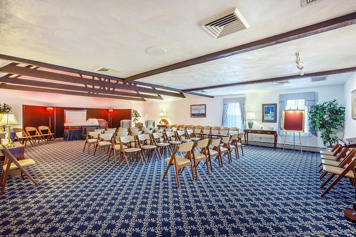 Conference room with rows of chairs facing a presentation area; blue patterned carpet, overhead beams.