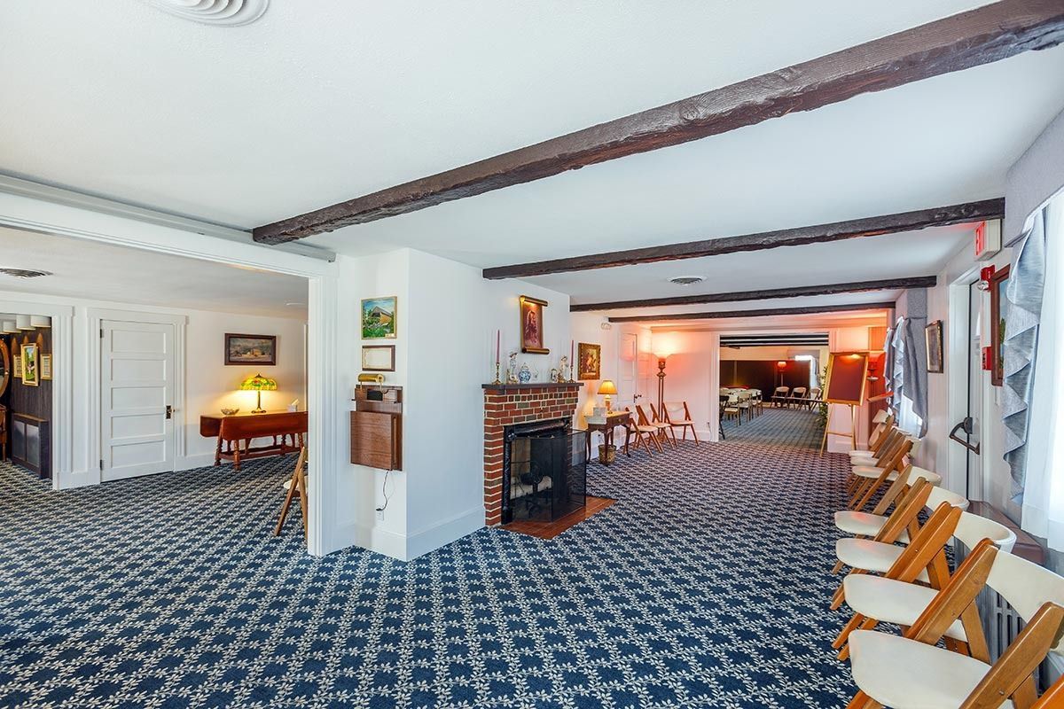 Interior of a room with a patterned carpet, fireplace, chairs, and doorways. Wooden beams cross the ceiling.