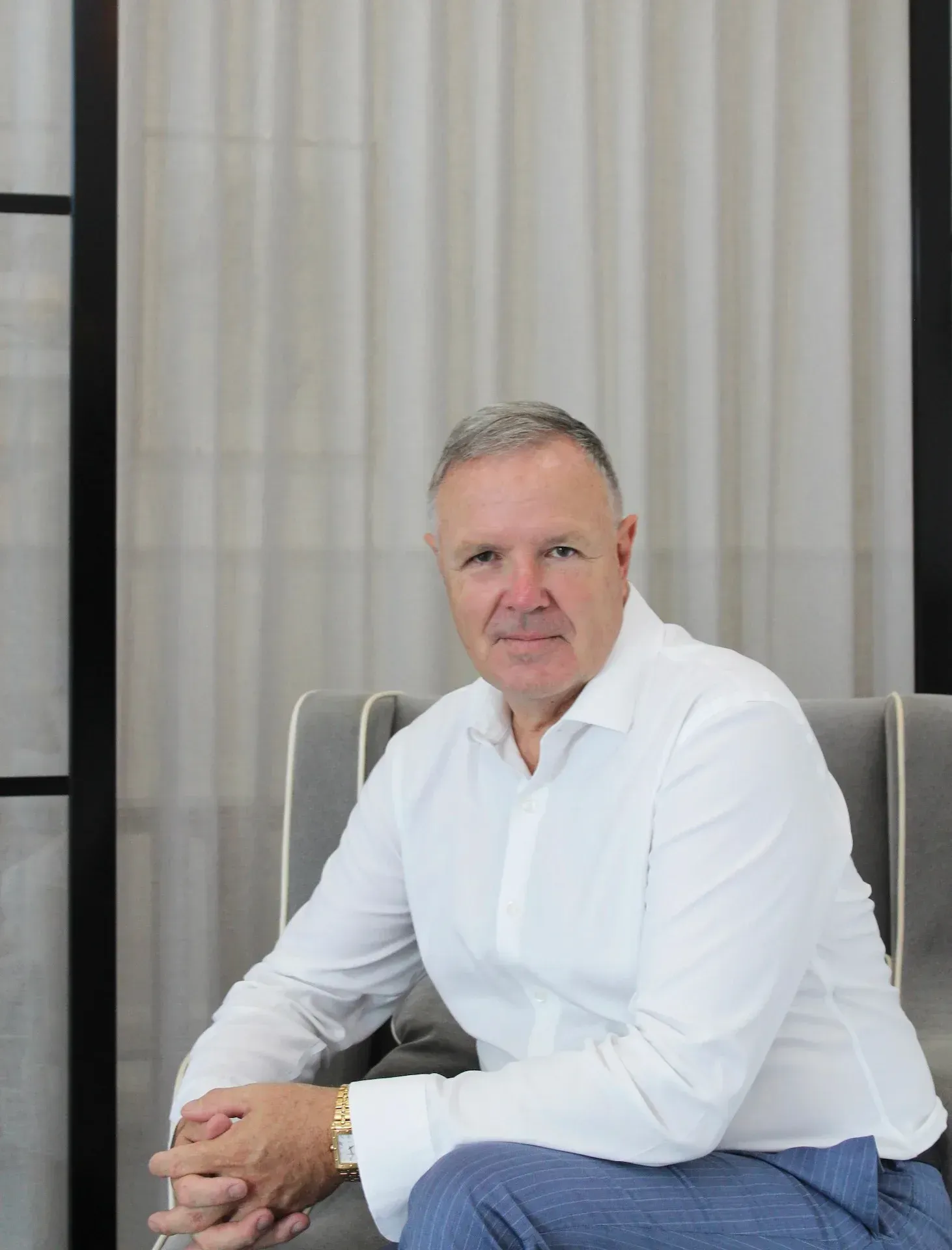 A man in a white shirt is sitting in a chair with his hands folded.