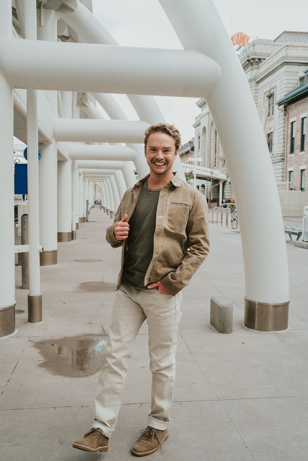 A man in a brown jacket and white pants is standing in front of a white pipe.