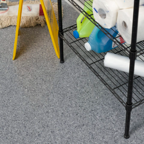 Wire storage shelf with paper towels, cleaning bottles, and a yellow mop bucket on a speckled floor