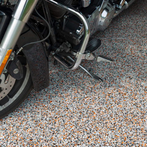 Close-up of a motorcycle parked on a speckled floor, showing the front wheel and engine guard