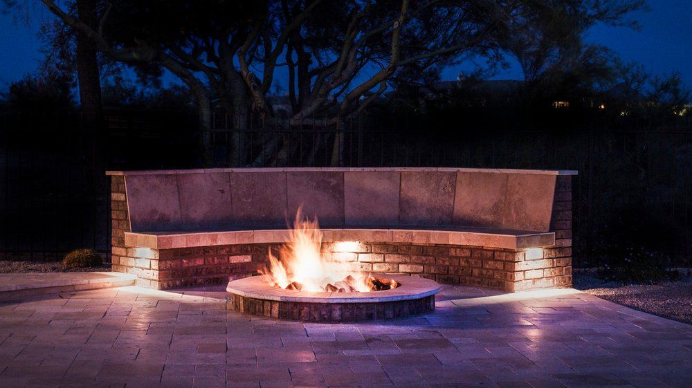 Fire pit with a stone bench at night. Flames in the center, surrounded by brick and stone, glowing lights.