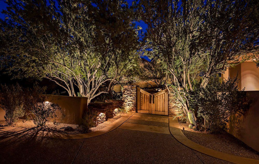 Night-lit driveway leads to wooden gate between trees, desert landscape, soft illumination.