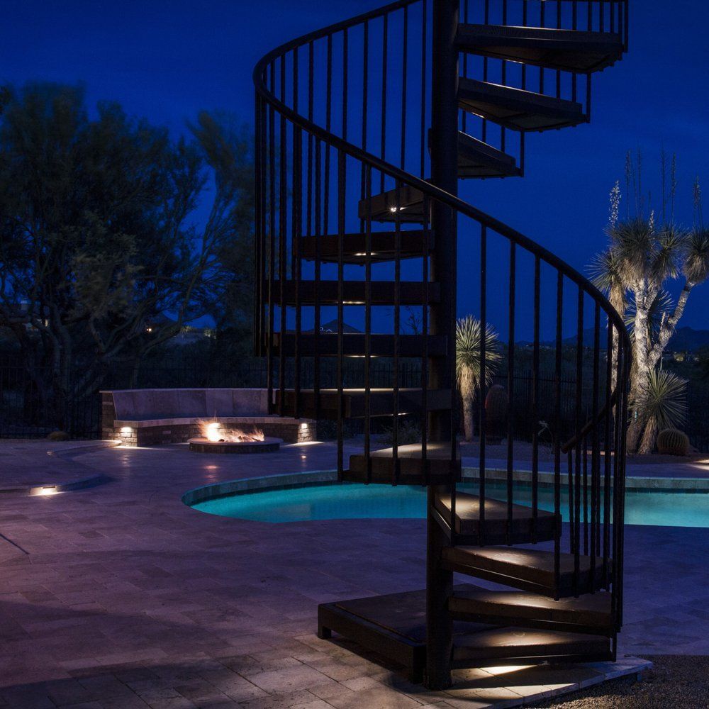 Black spiral staircase illuminated outdoors next to a pool at dusk.